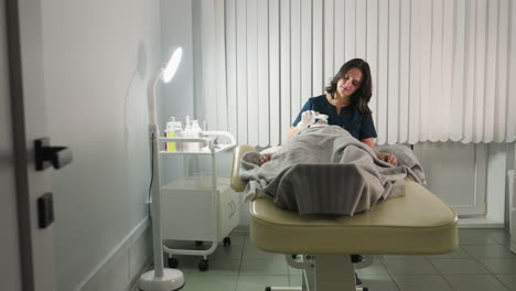 Skin Expert Using Electric Facial Brush Over Thick White Mask While Conversing With Relaxed Client Covered In Grey Blanket Under Bright Spa Lamp In Modern Minimalist Clinic Setting