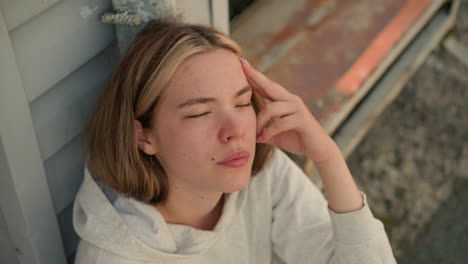 primer plano de una mujer joven descansando su mano por su cabeza, pareciendo perdida en profundos pensamientos, parpadea sus ojos, con una expresión tranquila e introspectiva, contra un fondo de pasos desgastados