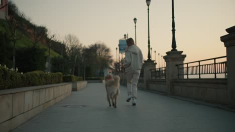 Vista-Trasera-De-Una-Joven-Rubia-Paseando-A-Su-Perro-En-El-Terraplén-Al-Amanecer