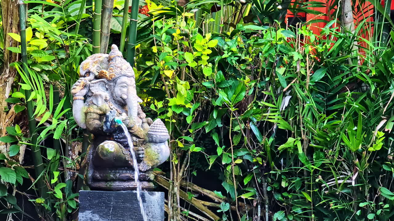 A traditional stone statue of the Hindu god Ganesha functions as a water fountain, surrounded by lush green tropical foliage and bamboo in a tranquil garden in Bali