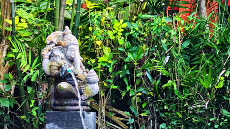 a traditional stone statue of the hindu god ganesha functions as a water fountain, surrounded by lush green tropical foliage and bamboo in a tranquil garden in bali