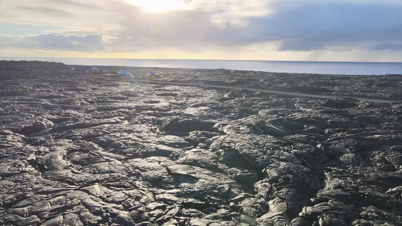 Dynamisk drönarbild som glider över en grusväg som slingrar sig genom ett vidsträckt lavafält, med ett litet samhälle badat i den milda morgonsolen, vilket visar upp den unika skönheten hos detta avlägsna landskap.