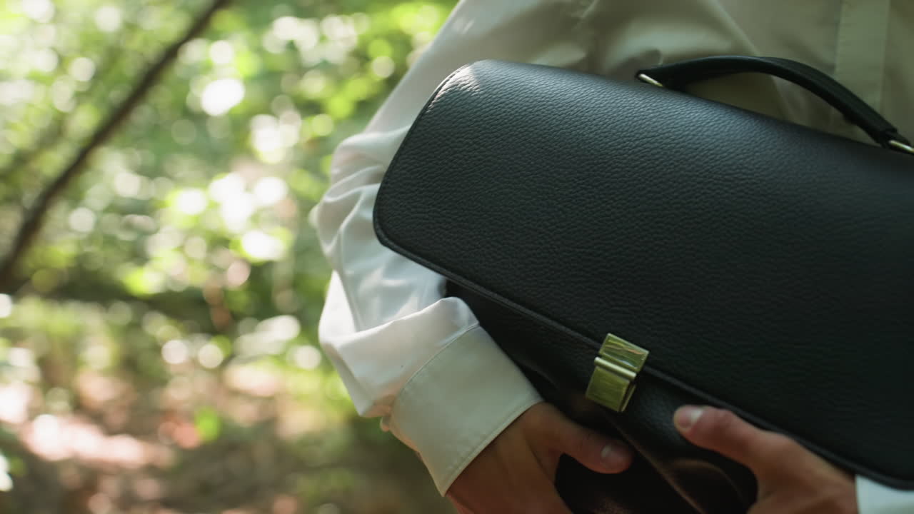 Biologist in white shirt carrying backpack tightly while bending carefully and walking through dense green forest under daylight, moving cautiously between branches and trees