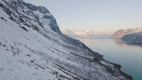 Aerial-along-steep-snow-covered-mountain-slope,-scenic-road-next-to-fjord