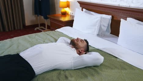 businessman rest at daytime. handsome man in white shirt and black trousers lying on bed in comfortable hotel. adult male relaxing during business trip.