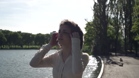 Portrait-of-a-fashionable-and-beautiful-Italian-model-walking-in-a-tranquil-park-near-a-lake,-wearing-a-transparent-shirt,-looking-around-and-having-fun-in-London,-United-Kingdom