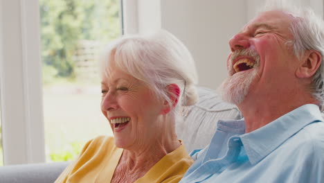 retired senior couple sitting on sofa at home watching tv and laughing together