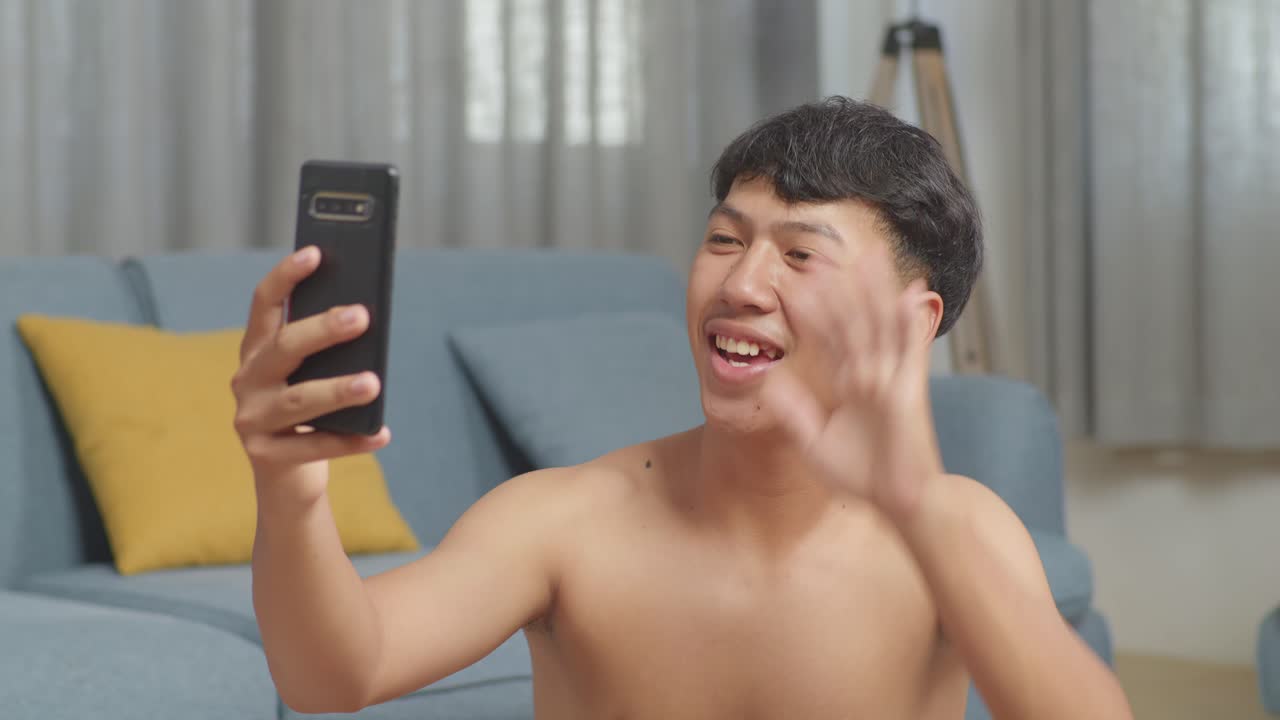 아시안 십대 소년은 집에서 셔츠를 벗고 운동한 후 덤벨과 함께 매트에 앉아 스마트폰으로 비디오 통화를 하는 동안 손을 흔들고 있습니다.