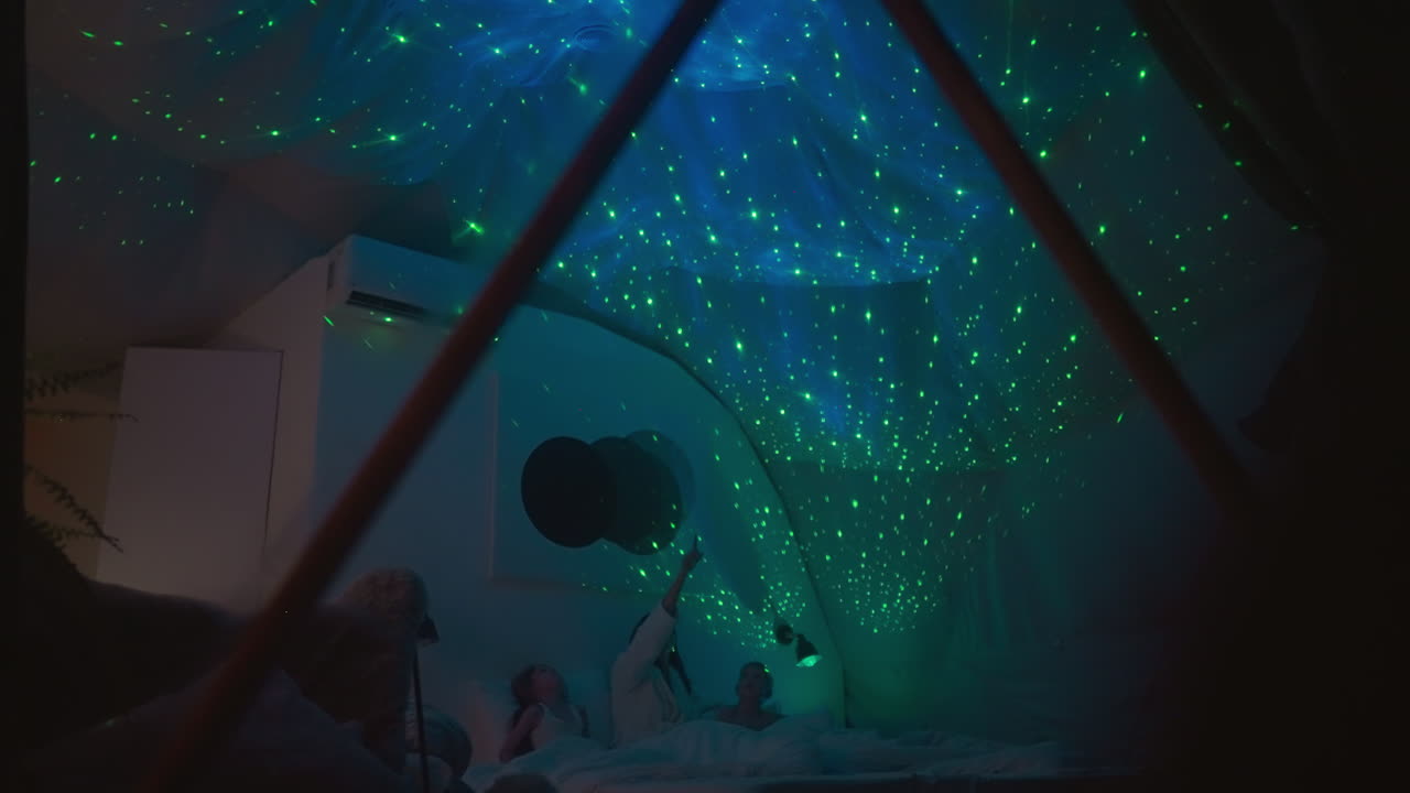 Little children with relative rest on large bed with reflection of night sky on ceiling. Glass glamping room with device creating soft light in contemporary room