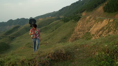 Asian-Male-Musician-Walks-with-Acoustic-Guitar-at-Green-Hills-Landscape,-Vintage-Clothing-and-Sunglasses