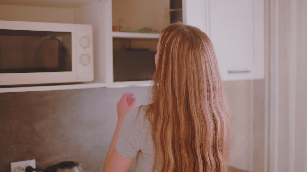 Uno studente si sveglia e va in cucina al mattino indossando dei pantaloncini, allungando la mano verso il contenitore del caffè nel mobile superiore per preparare la bevanda, illuminato da una calda luce naturale