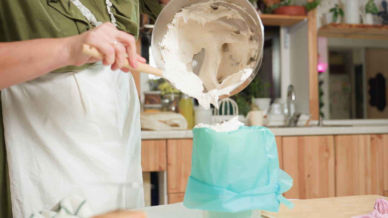 Baker in green gown uses wooden spatula to transfer thick creamy dough into piping bag held open by plastic container on kitchen table, preparing mixture for baking