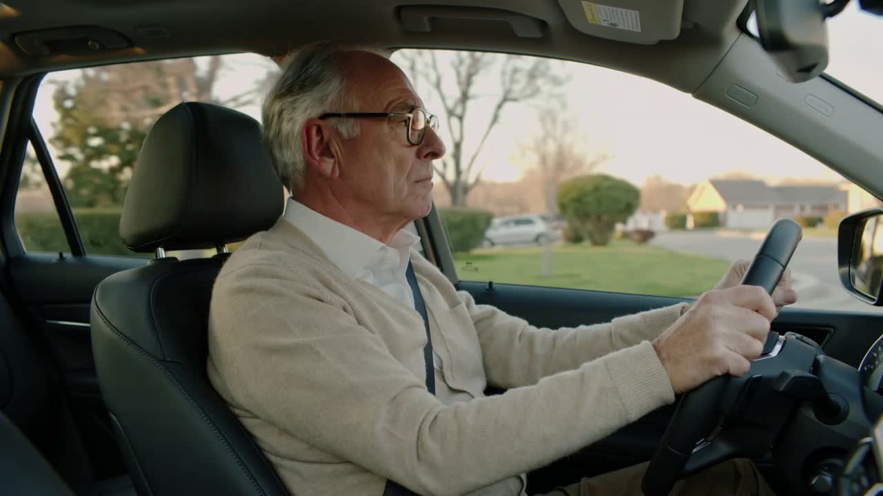 Driving senior man holding wheel as road curves through suburban street, wearing cardigan, seatbelt