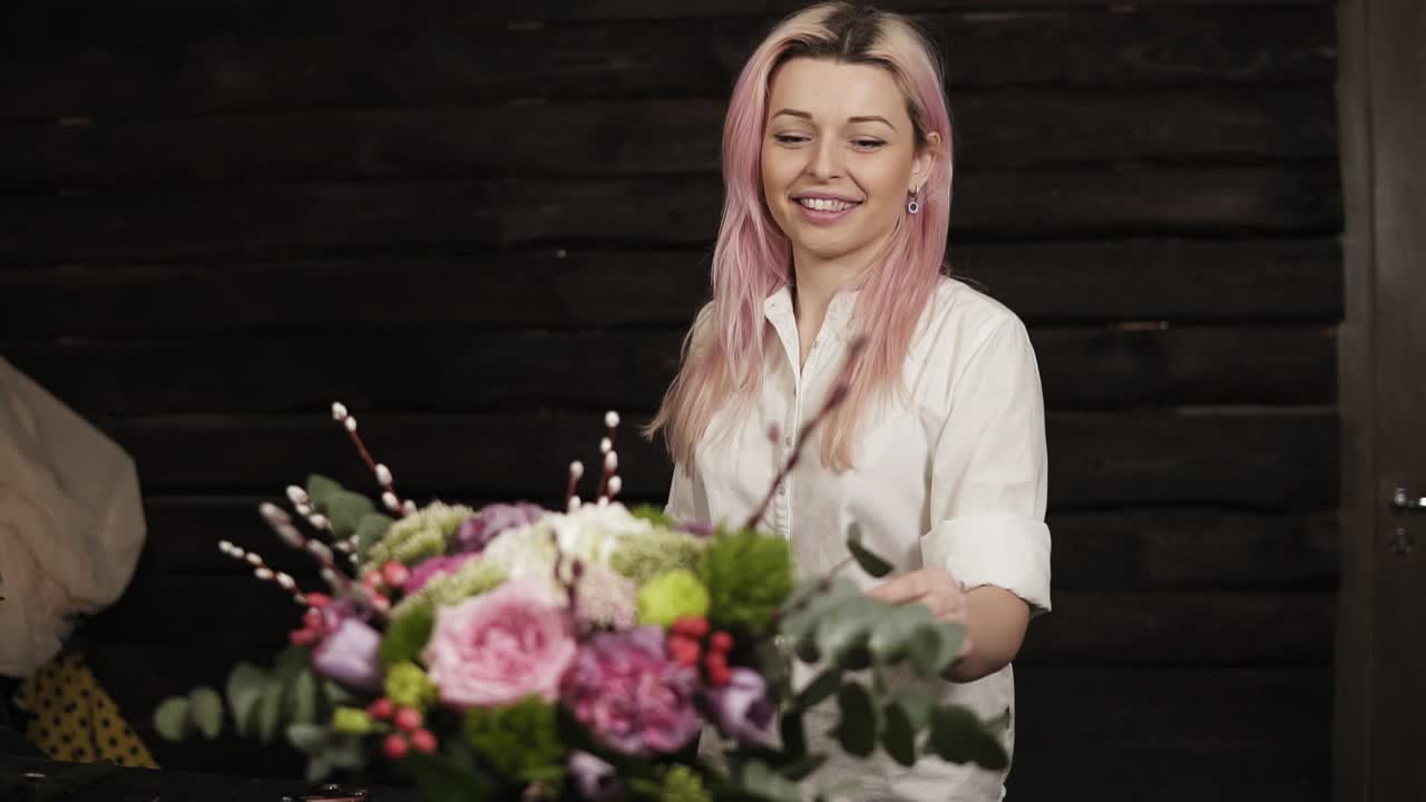 A beautiful girl with pink hair smiles, admires a stunning bouquet of flowers lying on the surface opposite her. The girl is a florist. Dark, modern studio