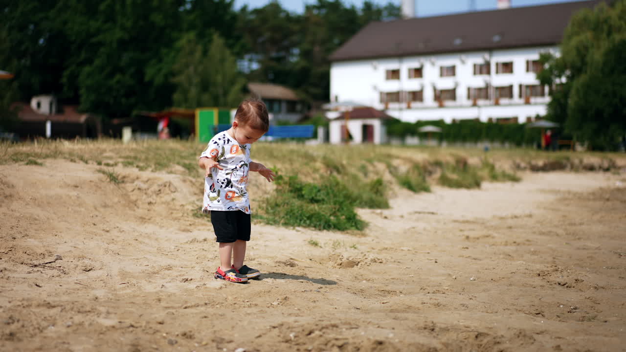 Happy baby boy stands in the sun on a sandy ground. Smiling kid runs looking at camera and around.