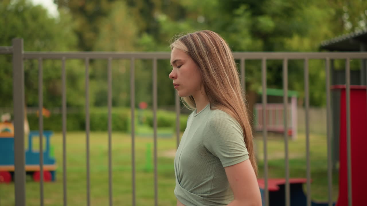 Side view of young woman walking thoughtfully along pathway beside iron fence, long blonde hair cascading down her back, with playground structures and green grass visible through fence