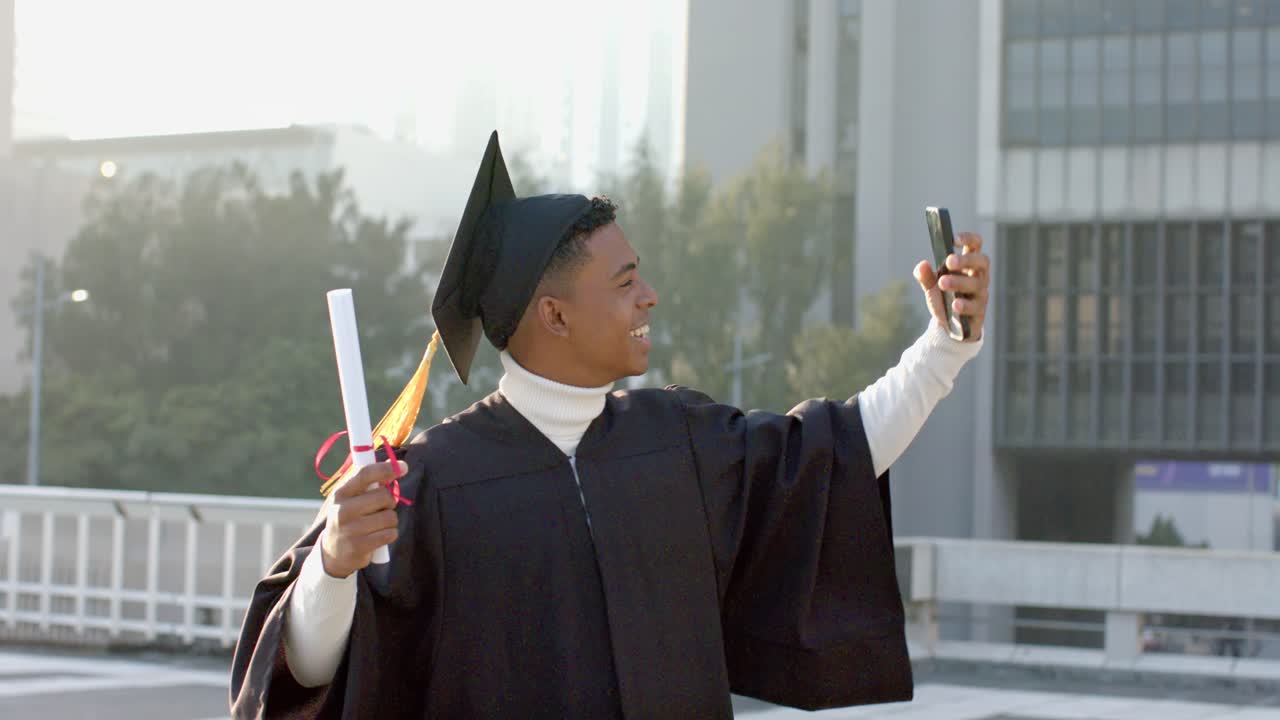 Male gowned graduate with diploma calling home, raising smartphone for selfie on terrace