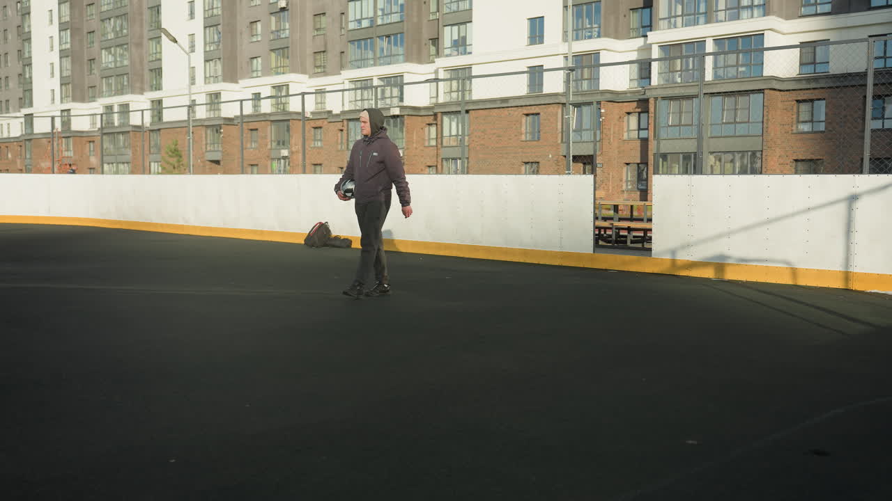 Individual walking on outdoor sports arena holding soccer ball, surrounded by urban architecture and bag close to the wall, with shadows and soft sunlight