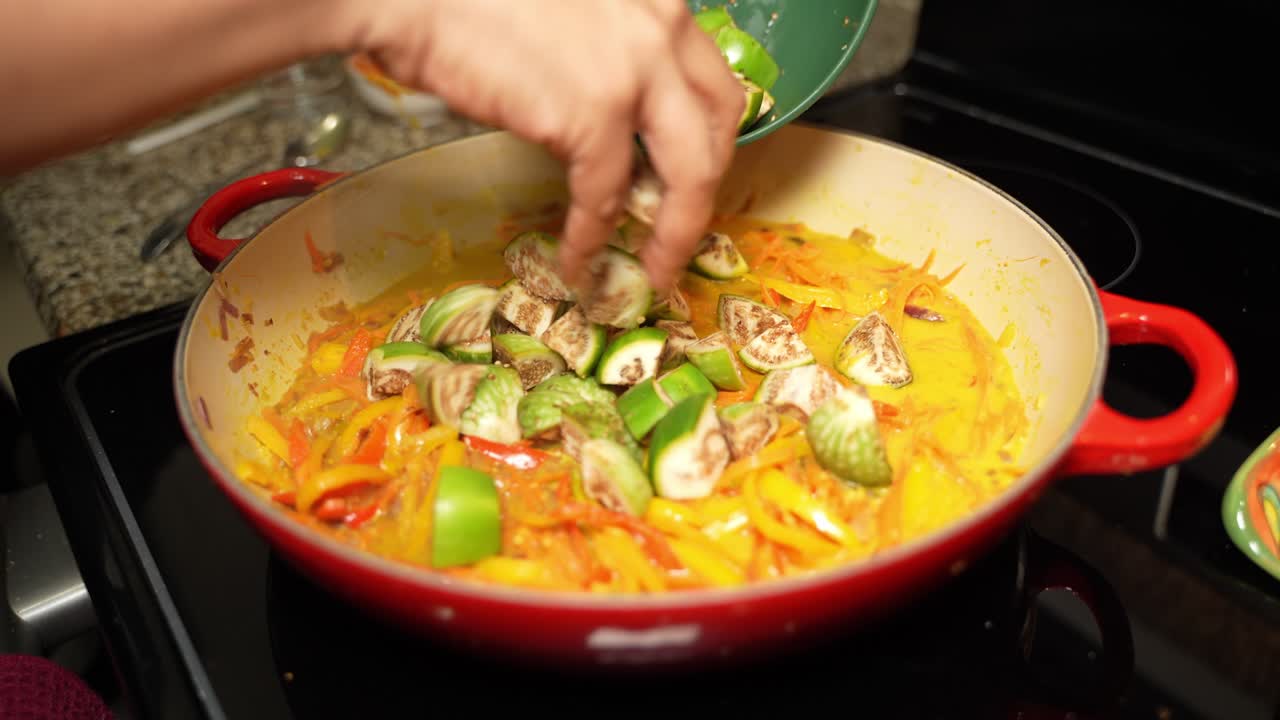 Hands of chef putting raw brinjals on frying pan with sliced bell paper. Veg recipe.