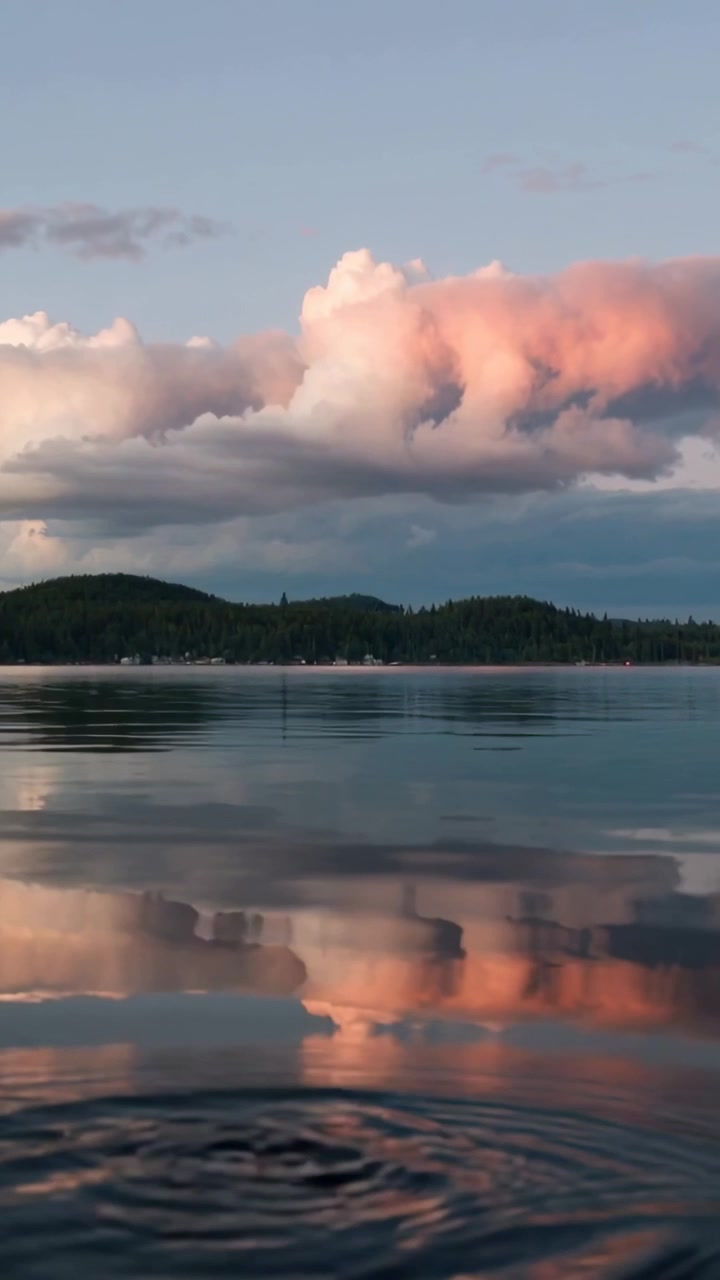 Vertikal video: Glimtende lyserøde skyer reflekteres i en rolig sø, et plask skaber ringe, der derefter forsvinder.