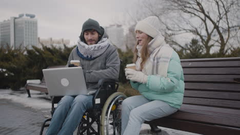 Behinderter-Mann-Im-Rollstuhl-Und-Sein-Freund-Schauen-Sich-Etwas-Lustiges-Auf-Dem-Laptop-An,-Während-Sie-Im-Winter-2-Im-Städtischen-Park-Kaffee-Zum-Mitnehmen-Trinken