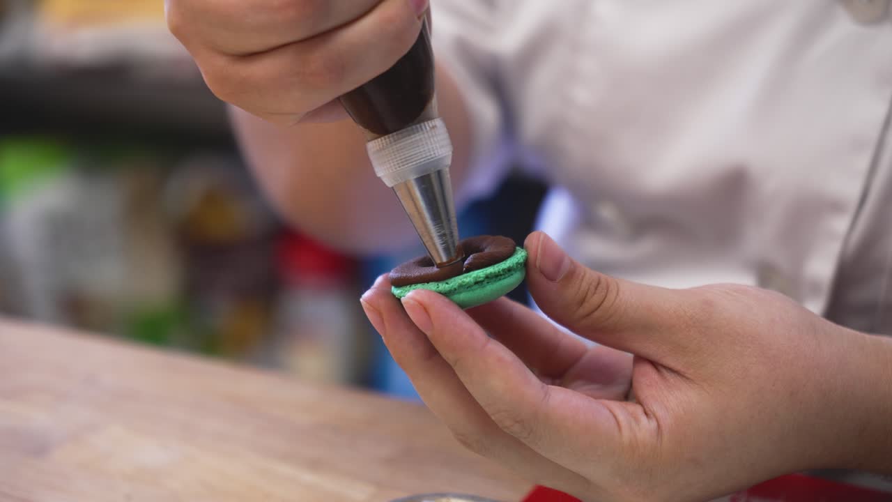 Pastry chef perfectly pipes chocolate cream filling onto French macaron cookie shell, slow motion close up 4K