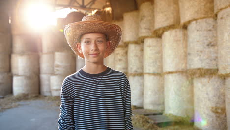 portret van een blanke tienerjongen met een hoed die naar de camera kijkt in een stal met hooivoorraden