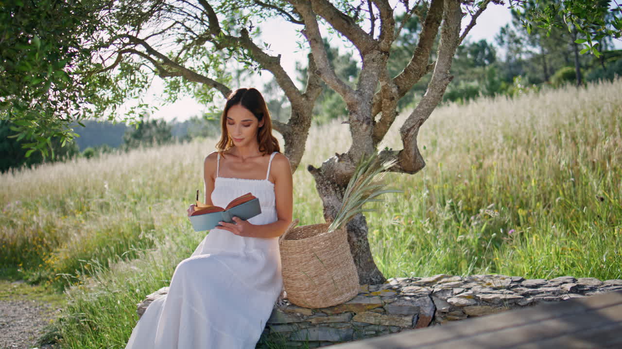 Dreamy girl opening novel sitting garden log. Woman enjoy book putting marks
