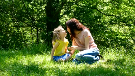 happy mother and little daughter playing at the park having picnic outdoors sitting on the green grass. young pretty brunette is talking to a cute girl.