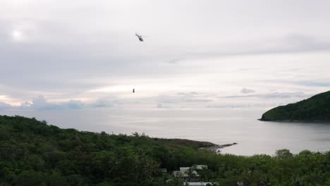 Helicopter-flying-away-with-supplies-hanging-from-rope-at-remote-island,-aerial