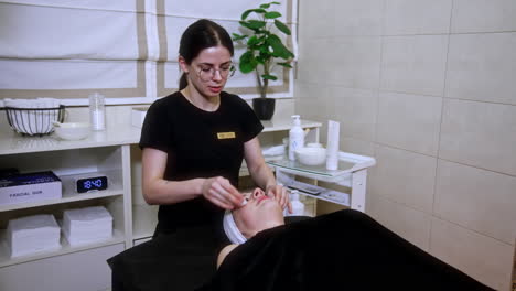 dark-haired female in glasses uses sponges to clean the client's face. beauty and spa procedures in the cosmetologist's cabinet.