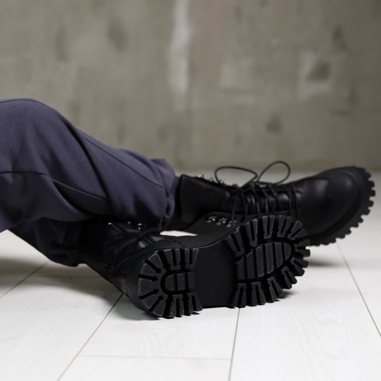 Model in grey sportive pants sitting on the white floor in studio. Female touching the tongues of black stylish army boots on her feet