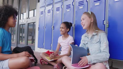Video-of-three-happy,-diverse-schoolgirls-talking,-sitting-by-lockers-in-school-corridor,-copy-space