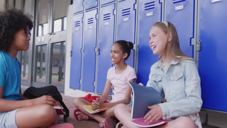 video of three happy, diverse schoolgirls talking, sitting by lockers in school corridor, copy space