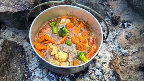 cocina de fuego de campamento, un delicioso estofado de horno de campamento con verduras hirviendo sobre las brasas en un pozo de fuego