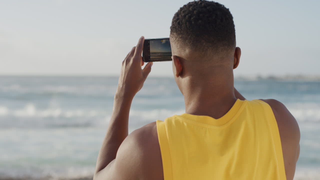 yoiung muscular african american man using smartphone taking photo of beautiful ocean seaside wearing yellow vest on beach enjoying vacation mobile sharing