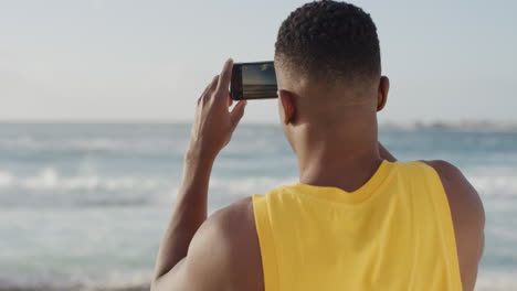 Junger,-Muskulöser-Afroamerikanischer-Mann,-Der-Mit-Seinem-Smartphone-Ein-Foto-Von-Der-Wunderschönen-Küste-Des-Ozeans-Macht-Und-Eine-Gelbe-Weste-Am-Strand-Trägt,-Während-Er-Den-Urlaub-Auf-Dem-Handy-Genießt