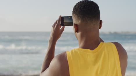 joven hombre afroamericano musculoso usando un teléfono inteligente tomando fotos de la hermosa orilla del océano con chaleco amarillo en la playa disfrutando de las vacaciones compartiendo móviles