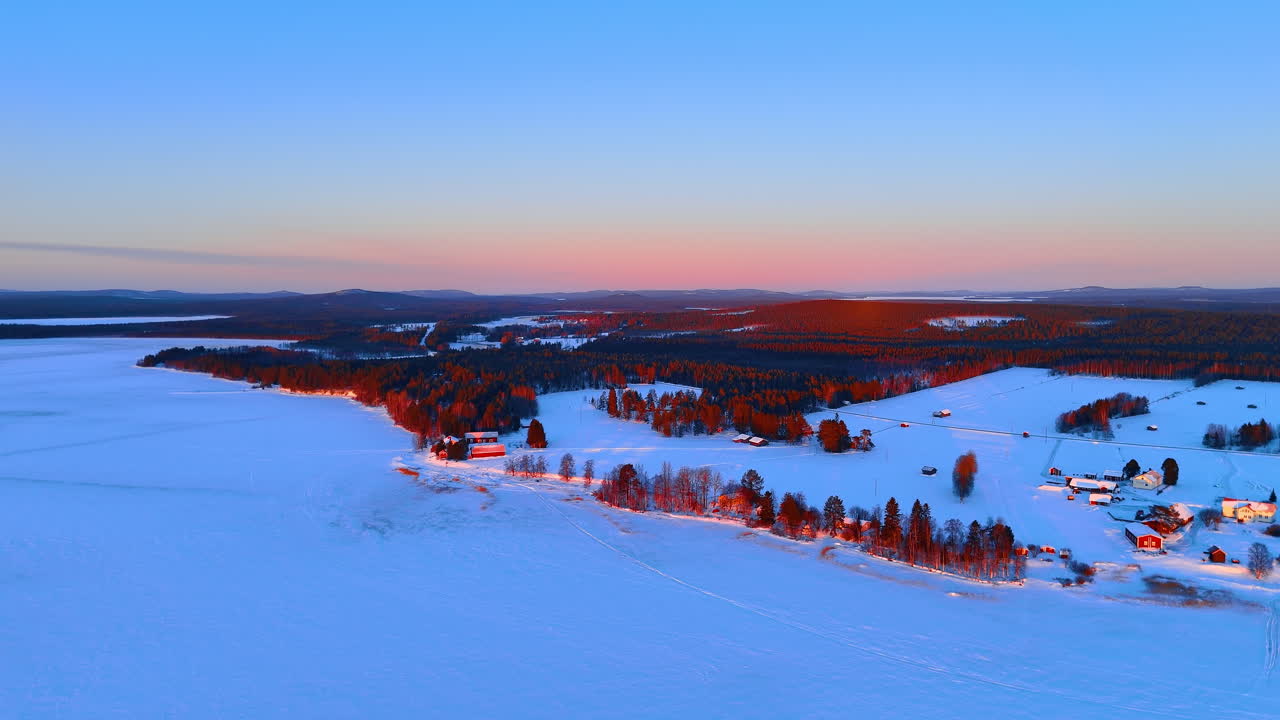 Snøkledd landsby og skog ved solnedgang. Fugleperspektiv over hus, trær og en frossen innsjø under gyllent solnedgangslys i Lappland.