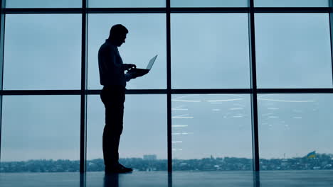 silhouette of a fit tall man standing at the window with laptop. man in glasses types on computer, closes gadget and walks along.