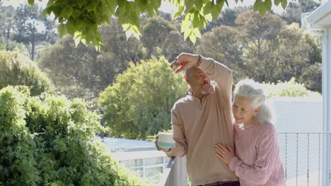 happy senior biracial couple embracing and drinking coffee at balcony at home, slow motion