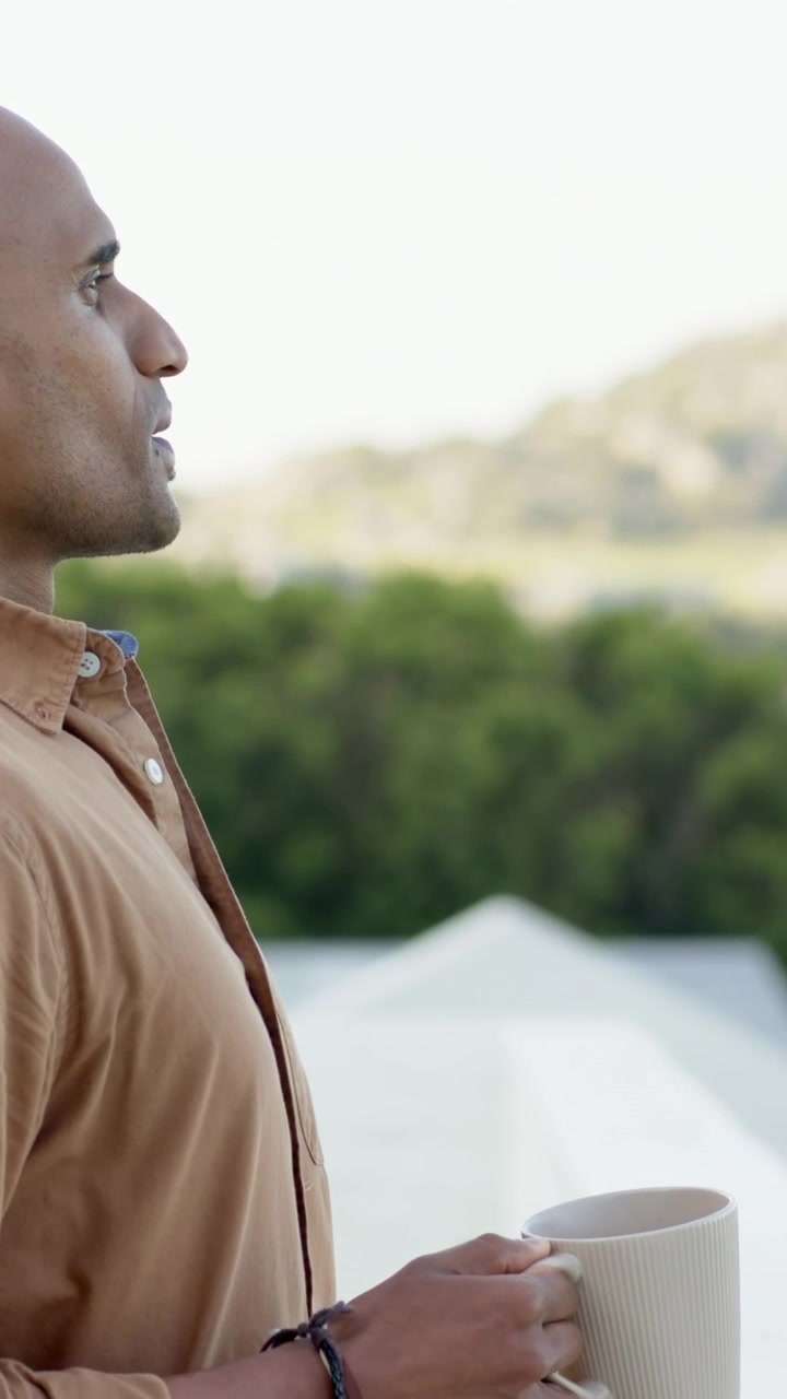 Vertical video: Pulling back camera, man holding mug on balcony, tan shirt, gazing, copy space