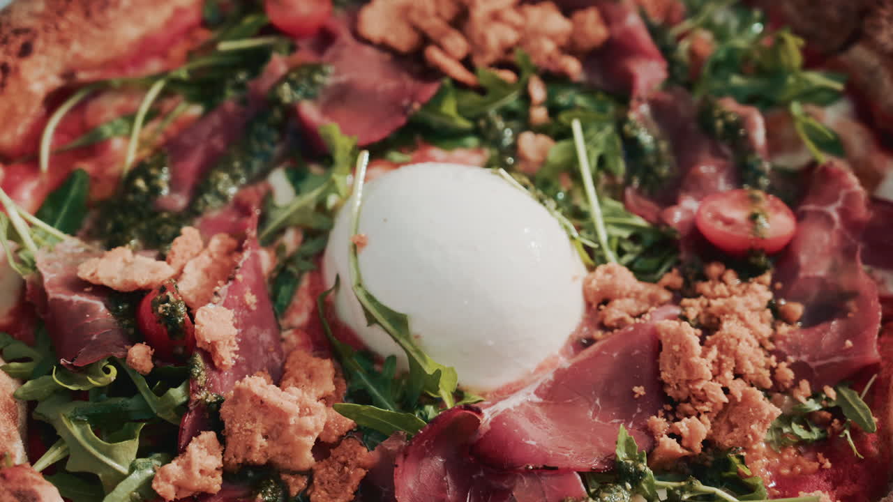 A beautifully shot overhead view of a gourmet pizza topped with burrata, prosciutto, cherry tomatoes, pesto, arugula, and crunchy crumbs