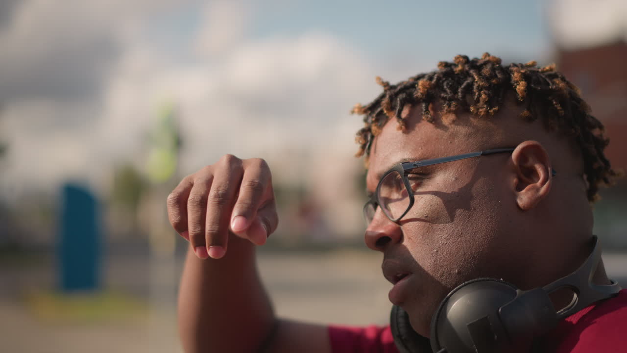 Nærbilde av svart DJ med hodetelefoner som justerer solbriller, varmt sollys, urban parkeringsplass som bakgrunn, flettet hår, reflekterende briller, fokusert uttrykk, filmatisk grunn dybdeskarphet.