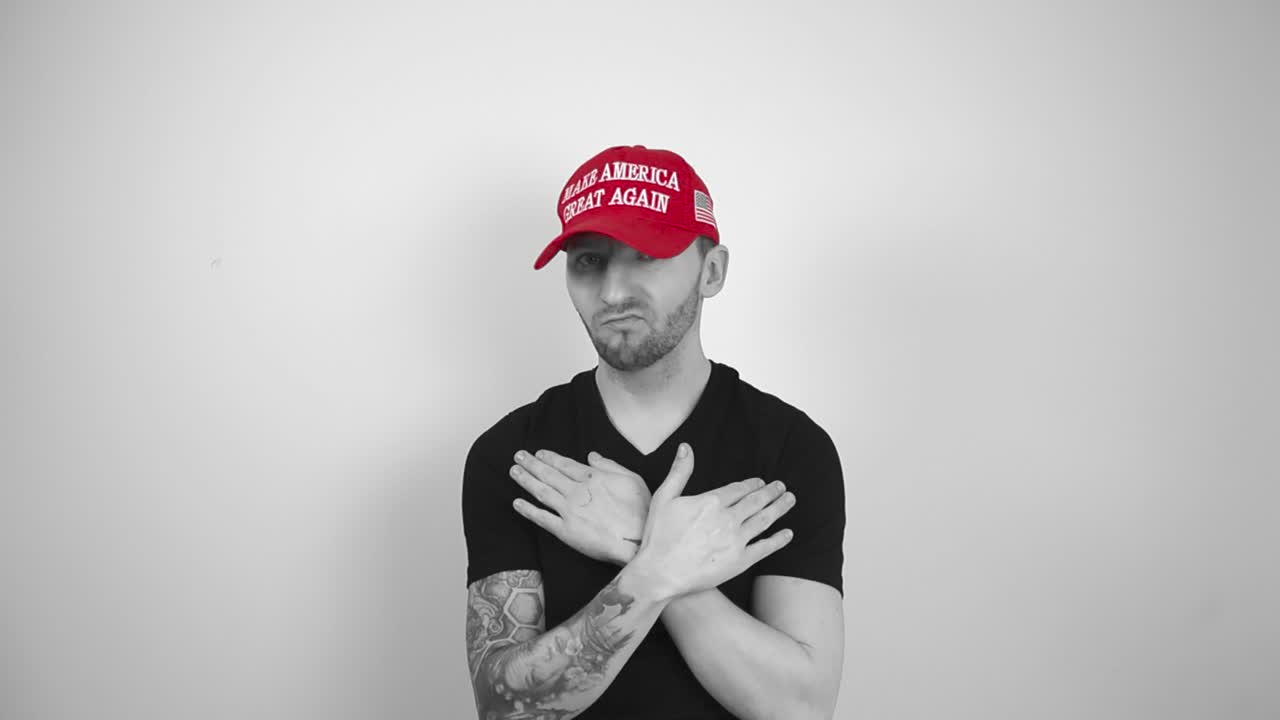 Grayscale footage of a tattooed man with a red MAGA Make America Great Again Trump hat moving hands around neck or neckline to show that he does not like something and you should stop. Man not happy.