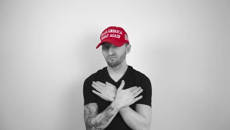 grayscale footage of a tattooed man with a red maga make america great again trump hat moving hands around neck or neckline to show that he does not like something and you should stop. man not happy.