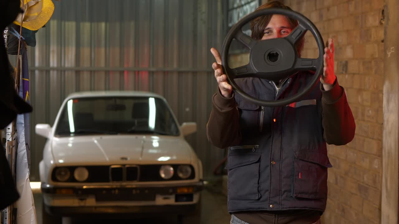 mecánico guapo joven y feliz jugando con el volante en el garaje con coche de época en el fondo