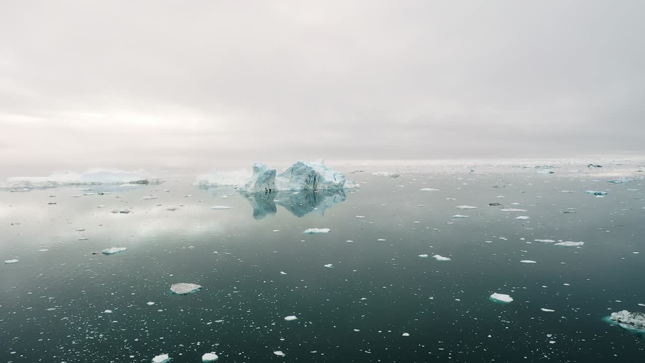Aerial drone view of icebergs floating on still Arctic sea under a cloudy sky in Greenland, showing peaceful reflections and serene beauty of the frozen polar environment