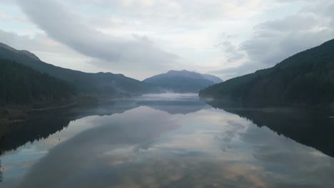 a long, calm lake reflecting the forested peaks, aerial rise