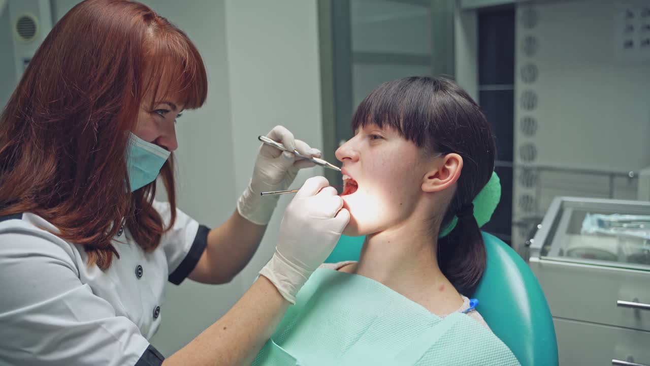 An experienced dentist in a white robe and mask examines the girl's teeth using a dental mirror and tweezers. Close-up.